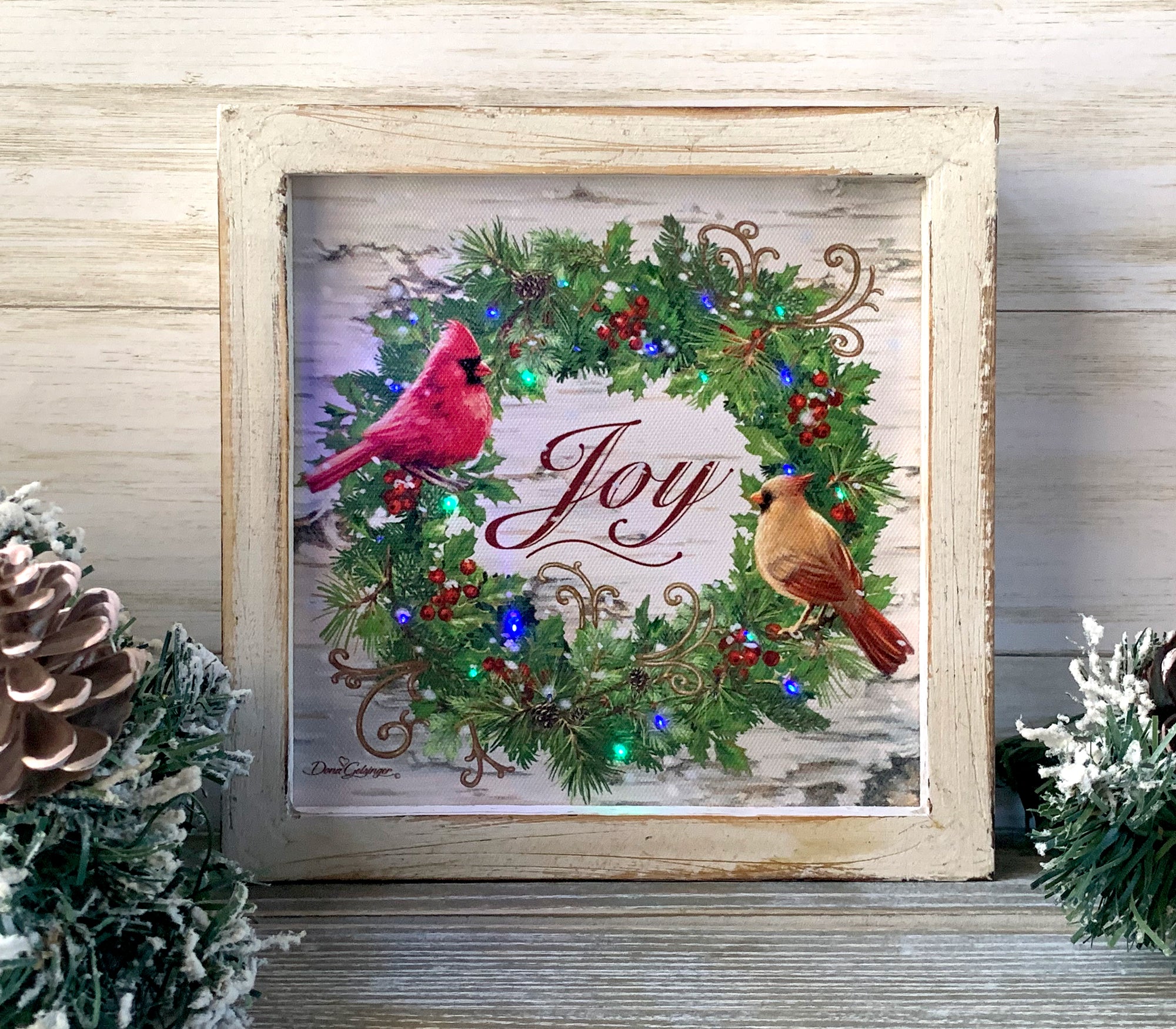 This stunning piece features two lovely cardinals perched on a magnificent wreath, complete with luscious berries, pinecones, and colorful lights that illuminate the scene with a warm glow.  The centerpiece of the wreath reads "joy"