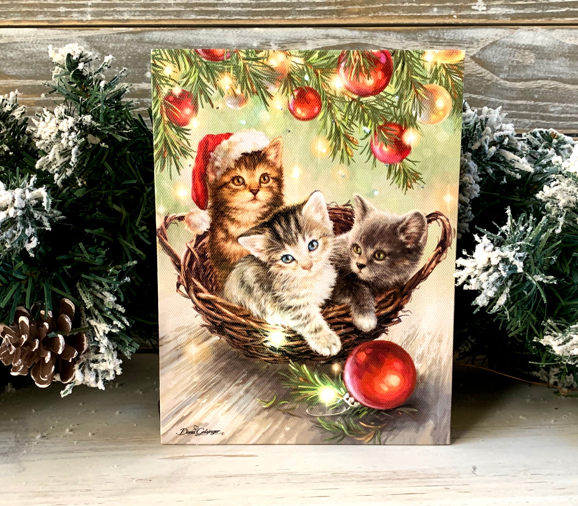 Three playful kittens snuggled up in a cozy woven basket, one of them donning a cute Santa hat, are sure to steal your heart.  As you gaze at the adorable scene, the mesmerizing bottom of a Christmas tree, adorned with twinkling lights and colorful bulbs, illuminates the canvas with a warm and inviting glow.