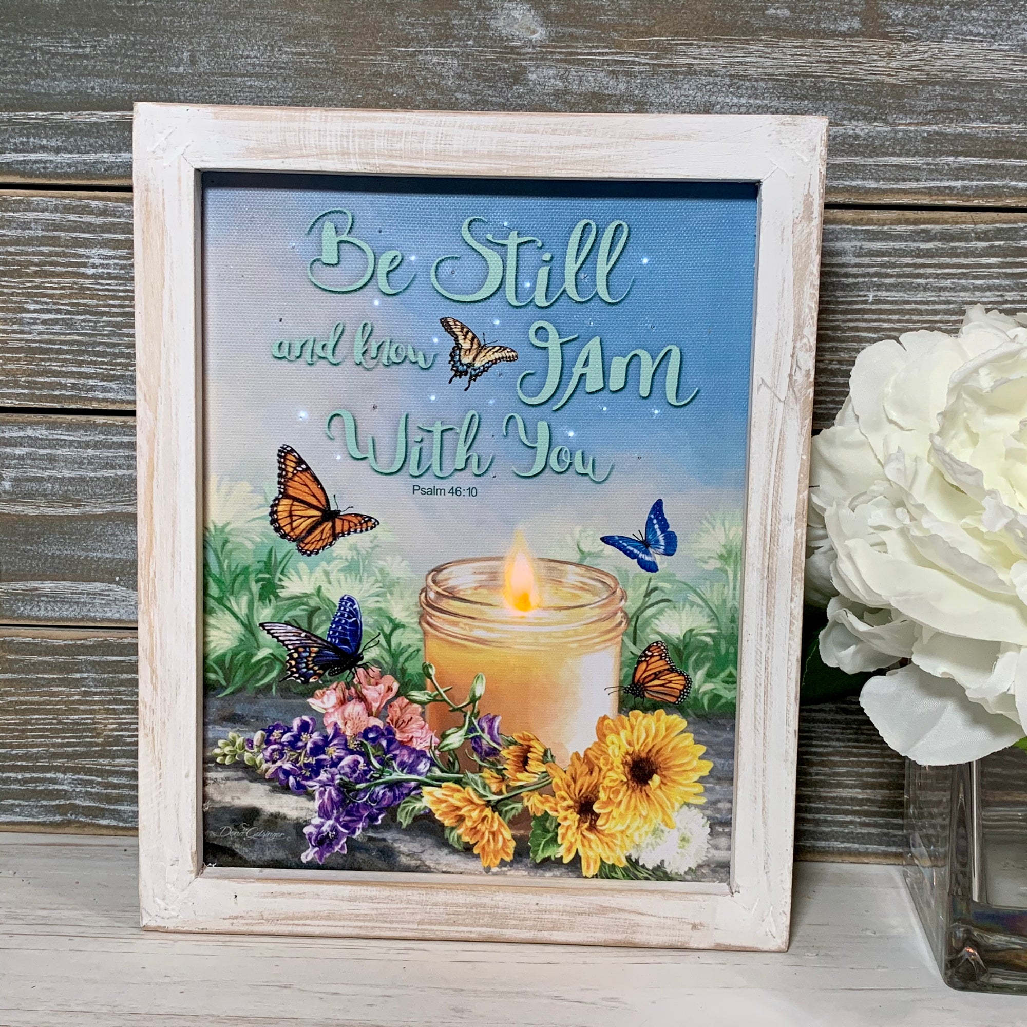 This beautiful candle, illuminated by gentle LED lights, casts a soft glow that adds an intimate and romantic ambiance to any room.  Surrounded by delicate flowers and fluttering butterflies, this shadow box is a symbol of new beginnings and the hope that love brings. And with the words of Psalm 46:10 written on the canvas, "Be still and know I am with you,"