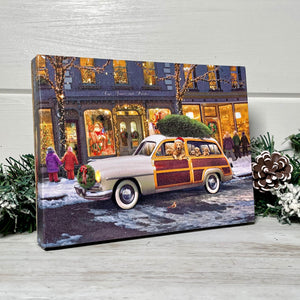  This stunning piece of art captures the essence of a romantic winter evening, where an old-time station wagon adorned with a wreath on the front and a fresh-cut tree tied to the roof is parked outside a row of charming storefronts.  As you gaze upon the scene, your eyes are drawn to the playful golden retriever and adorable puppies nestled inside the car.