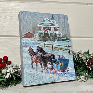 This stunning canvas captures the essence of a snowy wonderland with a beautiful scene of a horse-drawn sleigh ride. As the snow gently falls around them, a couple snuggles up close and enjoys the serene ride.  In the background, a charming Victorian-style house creates a picturesque backdrop to this idyllic winter scene. Another couple stands outside, admiring the beauty of the winter wonderland.