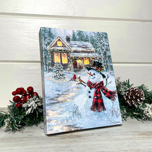 This enchanting piece features a charming snowman standing by a snow-covered path, leading the way to a cozy cabin nestled amongst towering pines. The snowman's top hat and plaid scarf add a touch of whimsy, while two cardinals perched on its wooden arm bring a delightful pop of color.