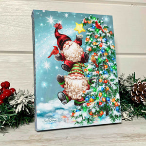 two adorable gnomes standing atop each other, spreading joy and cheer.  With a bright multicolored tree covered in snow and lights as their backdrop, these lovable gnomes are joined by a charming cardinal perched on the hand of the gnome on top.