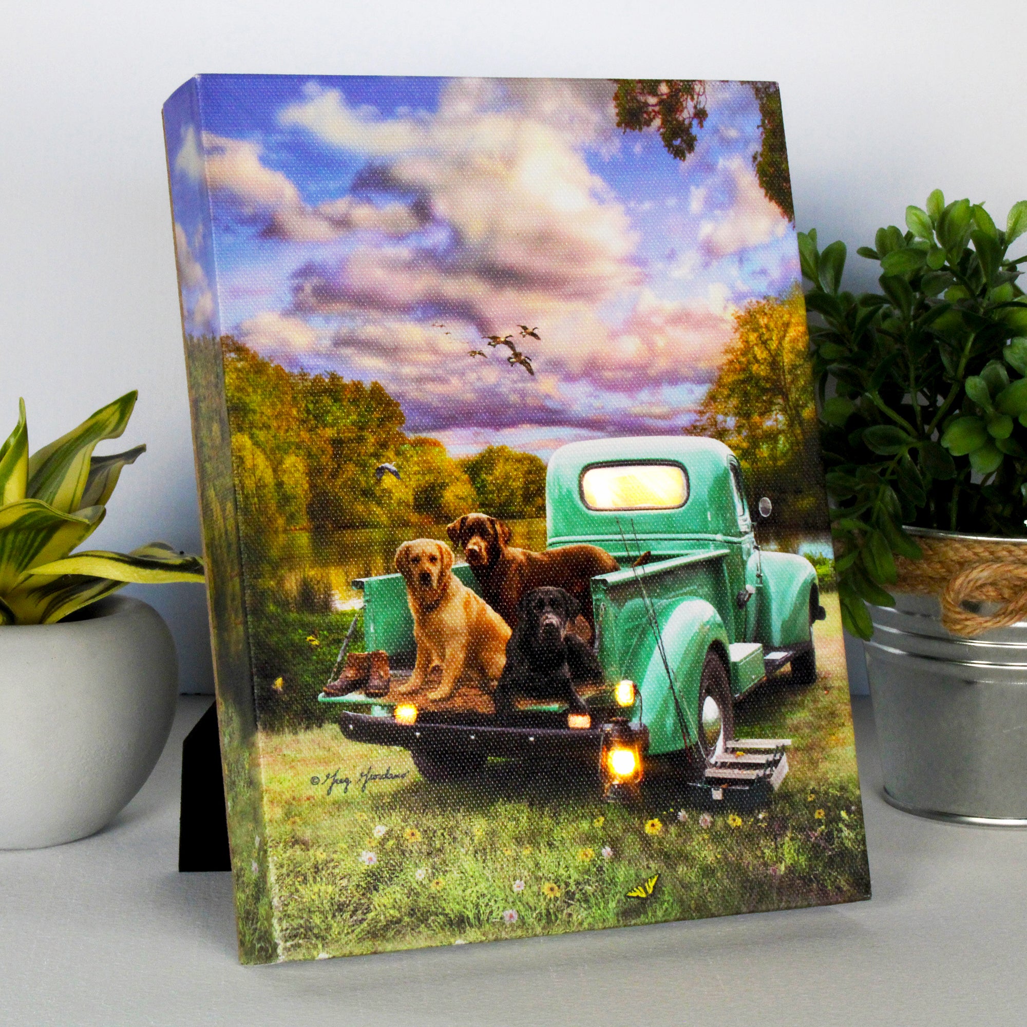 This beautiful canvas depicts an idyllic scene by the lake, where an old green truck is parked on the edge of the water, surrounded by nature's serene beauty. Three loyal Labrador retrievers sit proudly in the truck bed, eagerly anticipating the day's fishing trip. In the background, a flock of ducks flies overhead, adding a touch of whimsy to this charming scene.