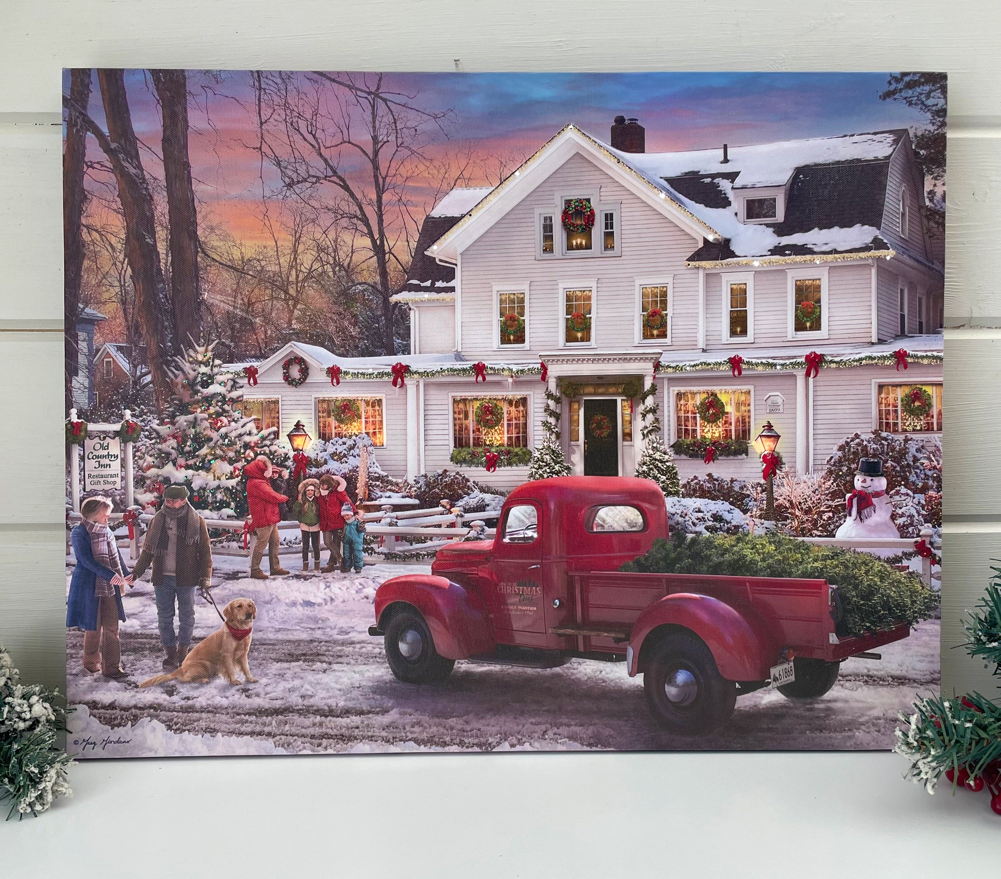 an old red truck sits parked in front of a picturesque white country inn. The truck bed holds a fresh cut tree, ready to be decorated and enjoyed by the fireplace inside.  The inn is adorned with wreaths and lights, setting the scene for a magical holiday season. A snowman stands tall in the front yard.