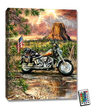 Featuring a stunning image of a Harley Davidson motorcycle, complete with fiery flames and the iconic American flag adorning the back, this piece of art captures the spirit of the open road and the thrill of the ride. The motorcycle sits on a dusty dirt road, with a backdrop of lush forest and a towering plateau rising high above the landscape.