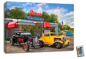 This stunning piece features two classic hot rods, perfectly polished and primed, as they await their turn to roll out onto the open road. The backdrop? A charming old-fashioned diner, straight out of the heyday of Route 66.