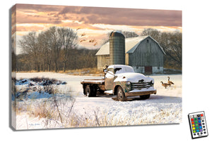 a vintage truck sitting peacefully in a snowy field, perfectly capturing the serene ambiance of a winter wonderland.  As you gaze upon the artwork, you'll notice two graceful geese standing nearby, adding a touch of wildlife charm to the scene. Above the barn in the background, a flock of geese can be seen flying gracefully through the crisp, blue sky.
