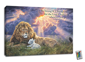 Behold a majestic lion, his mane flowing in the wind, lying in a field next to a gentle lamb. The sun streams through the clouds above, casting a warm glow on this beautiful scene.  With the inspiring words of Joshua 1:9 written on the canvas
