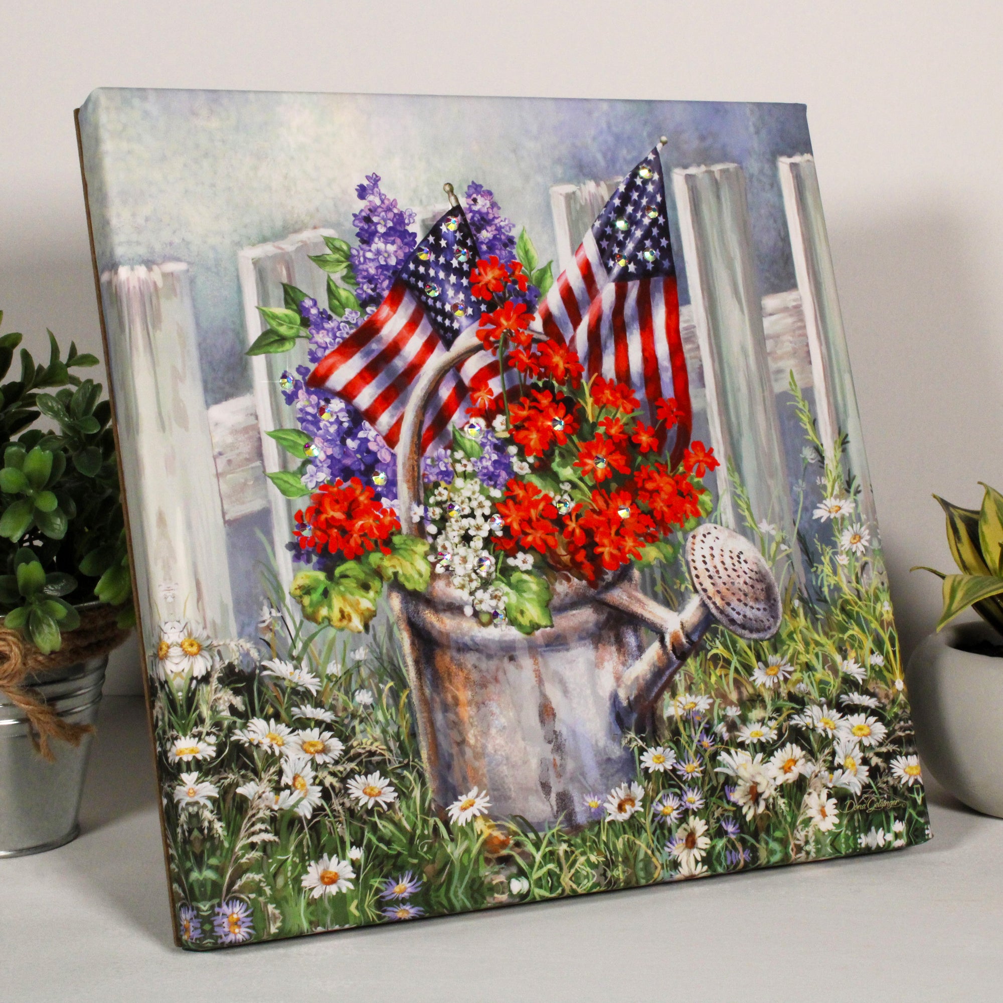 This stunning piece features a vibrant watering can nestled among a bed of colorful flowers, framed by a classic white picket fence. Adorned with brilliant crystals, this print truly sparkles with beauty and patriotism.  The vibrant hues of red, white, and blue are proudly displayed in the stunning flowers bursting forth from the watering can, while two American flags flutter from the watering can.