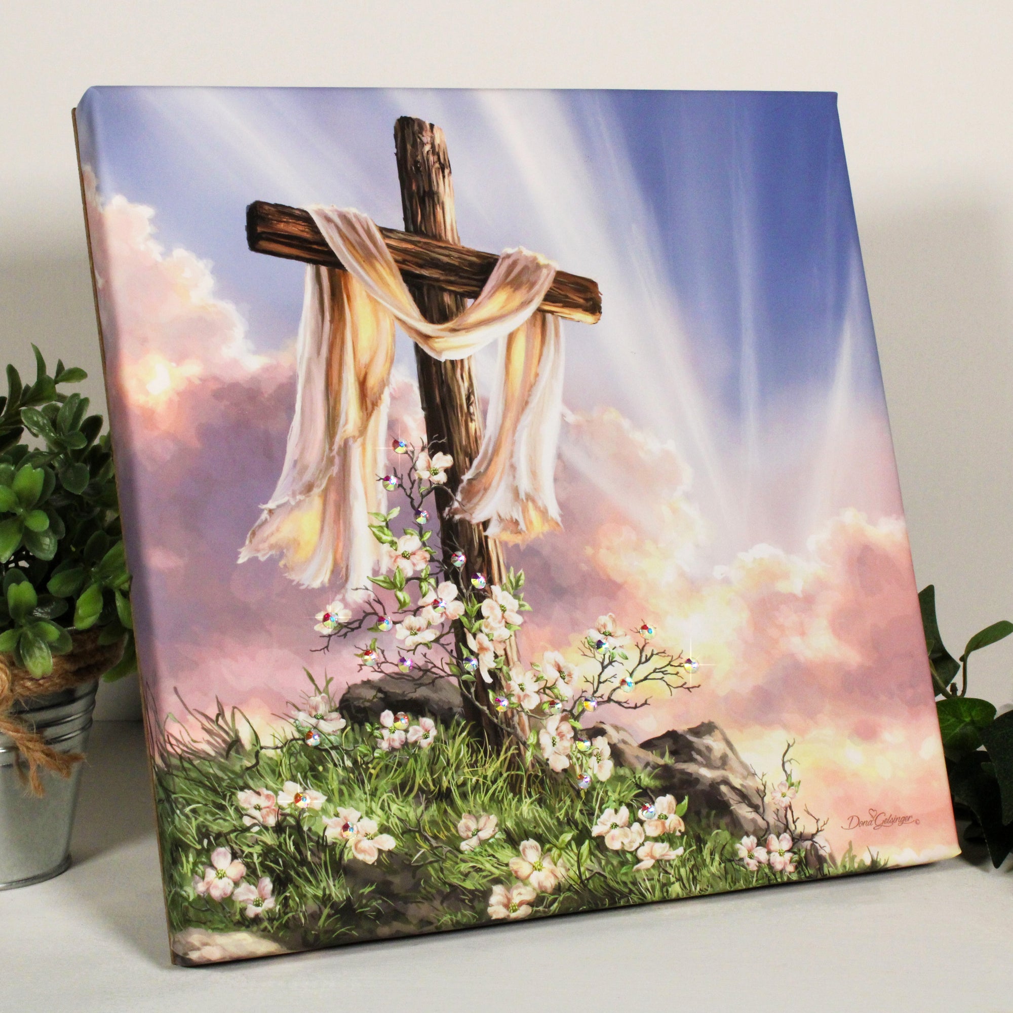 This stunning piece of art captures the essence of the resurrection with a beautiful cross perched atop a hill, draped in cloth, and adorned with white flowers. As the sun's rays gently stream through the scene, the message "he is risen" is boldly emblazoned on the canvas.