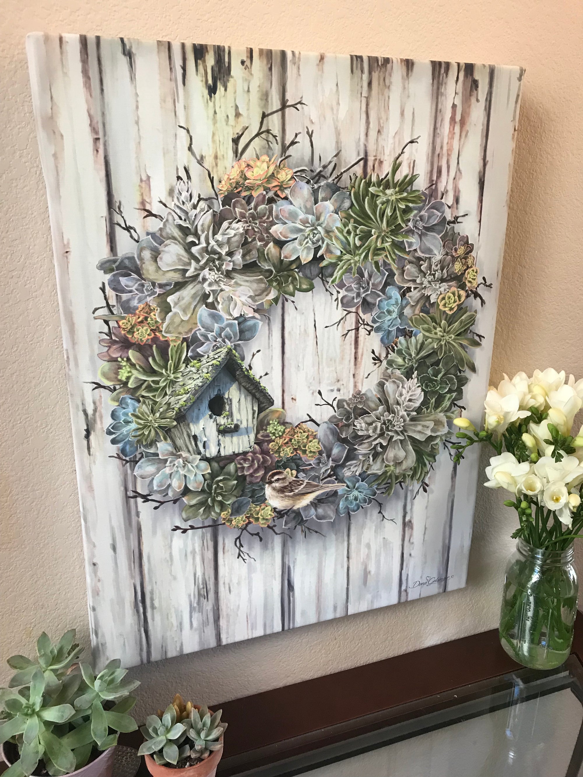This exquisite canvas features a wreath filled with an array of multicolored green, blue, yellow, and purple succulents, creating a vibrant and captivating display.  As you gaze upon this masterpiece, you'll notice a charming bird house nestled among the succulents, adding a whimsical touch to the scene.